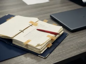 Opengeslagen certificeringmap met ezelsoren en sticky notes op een kantoorbureau, rode pen op onvolledige checklistpagina's, laptop op achtergrond.
